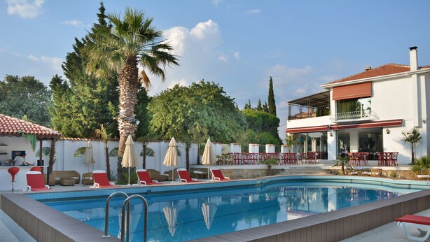 Una piscina con sillones de descanso y sombrillas, rodeada de follaje y un edificio de dos plantas con terraza.