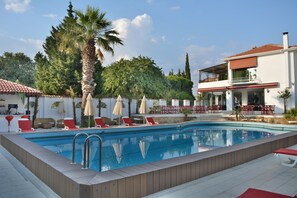Ein Swimmingpool mit Liegestühlen und Sonnenschirmen, umgeben von Grünpflanzen und einem zweistöckigen Gebäude mit Außenbereich.