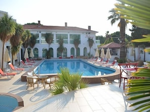 Un área de piscina con sillones de descanso y sombrillas, un edificio con arcos y palmeras.