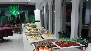 Montaje de bufé con diversos alimentos en bandejas y fuentes, dispuesto en una sala amplia con suelo de baldosas y ventanas arqueadas.