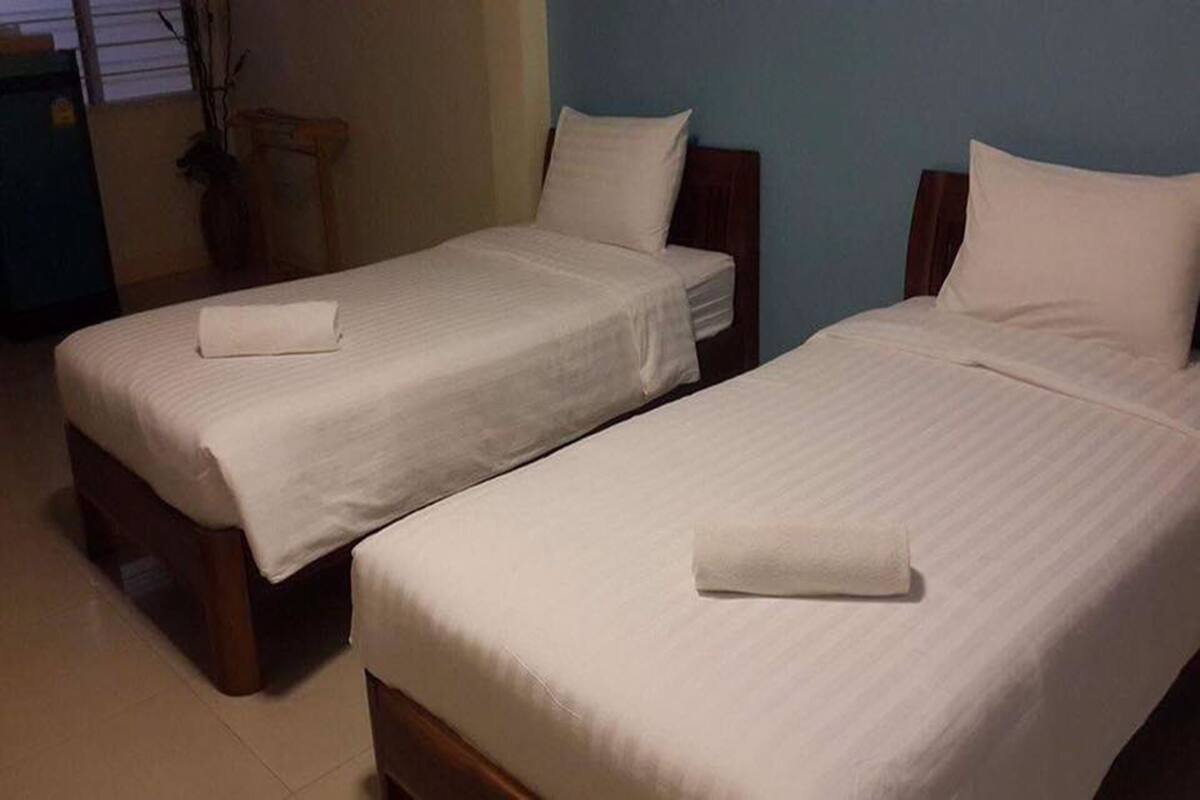 Dos camas individuales con sábanas y almohadas blancas, cabeceros de madera y una mesita con un florero en una habitación con paredes azul claro.