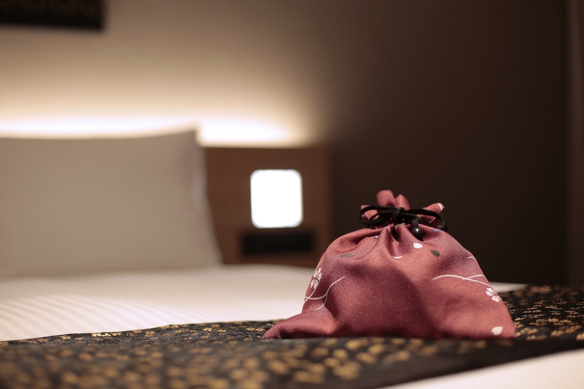 Una cama con una colcha estampada, una pequeña bolsa roja sobre la cama y una mesita de noche con una lámpara.