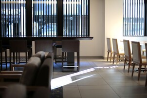 A modern interior with a row of chairs and tables, large windows with blinds, and a view of the outside.