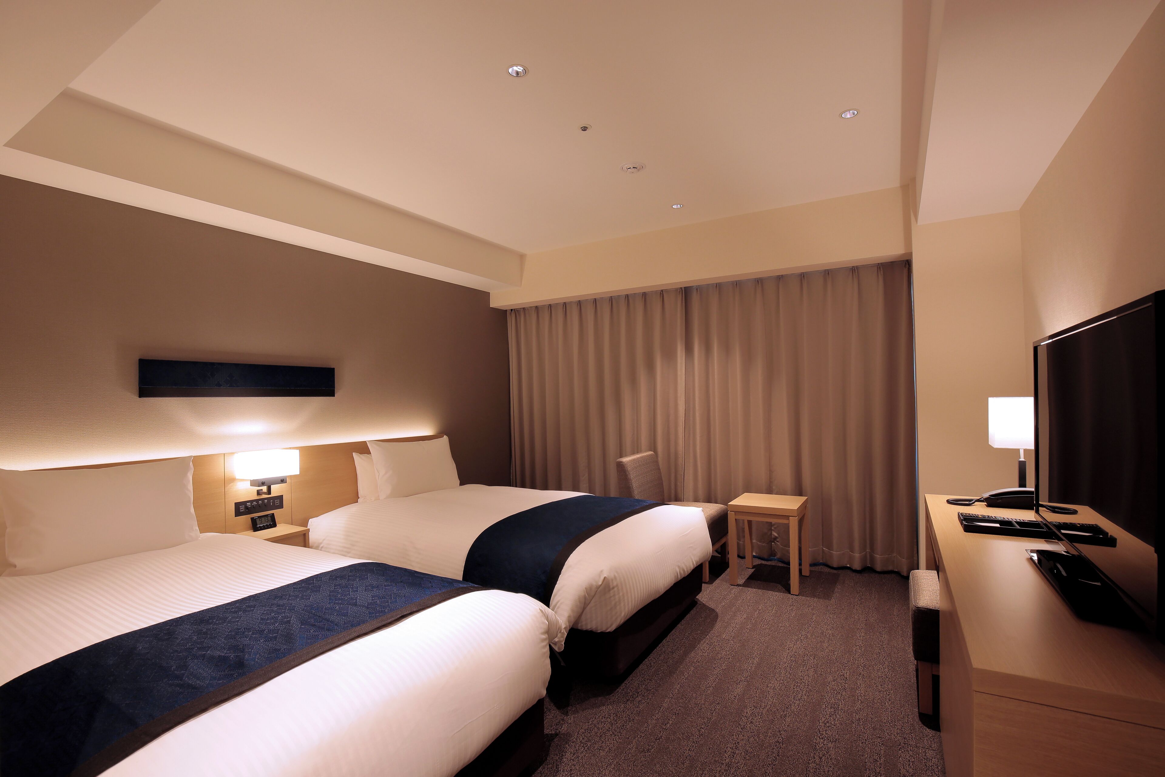 A hotel room with two beds, a desk, a chair, a TV, and a lamp.
