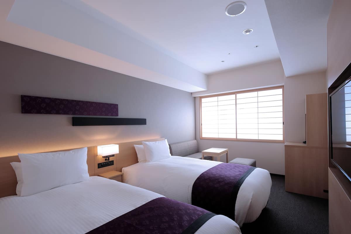 Una habitación de hotel con dos camas, una mesita, un televisor y una ventana con persianas.