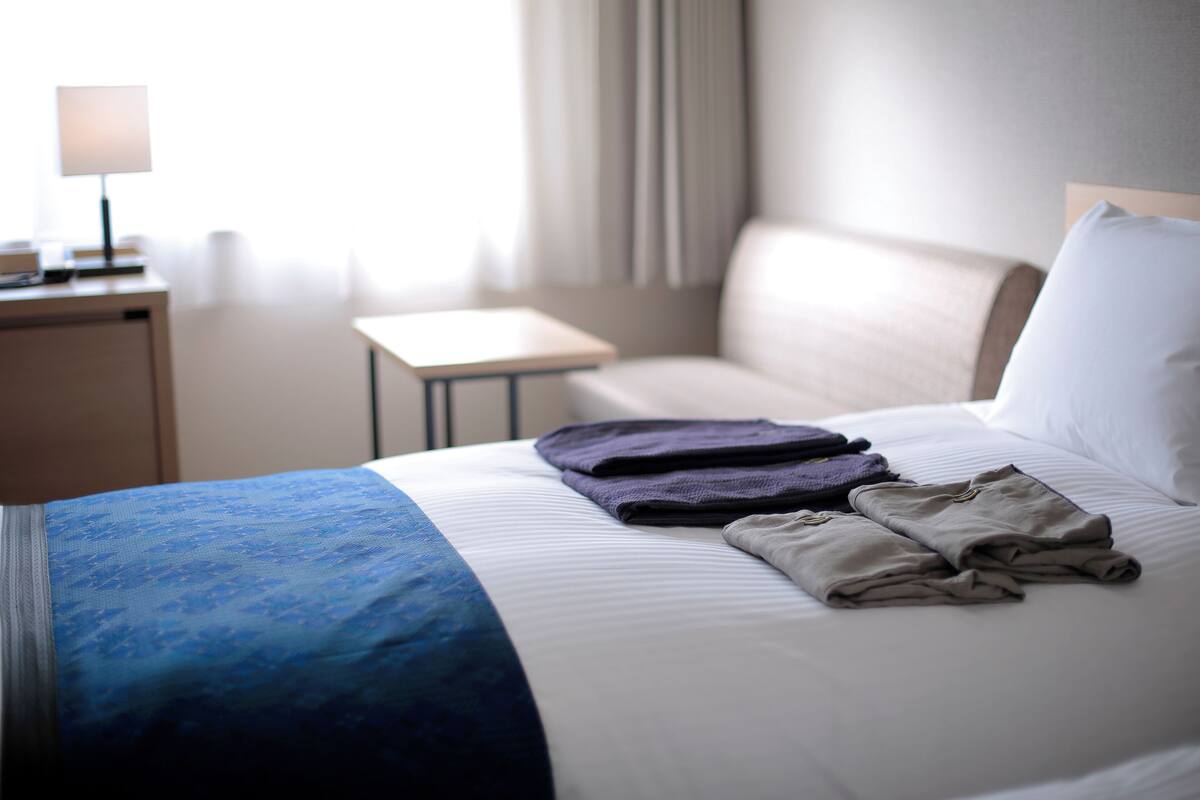Una cama bien hecha con una colcha azul y blanca, una manta gris doblada y dos toallas moradas dobladas.