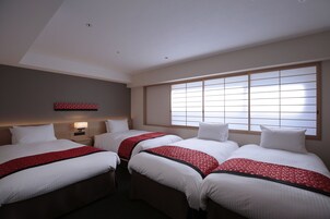 Habitación de hotel con tres camas, un ventanal con persianas, un televisor de pared y una mesita de noche con lámpara.