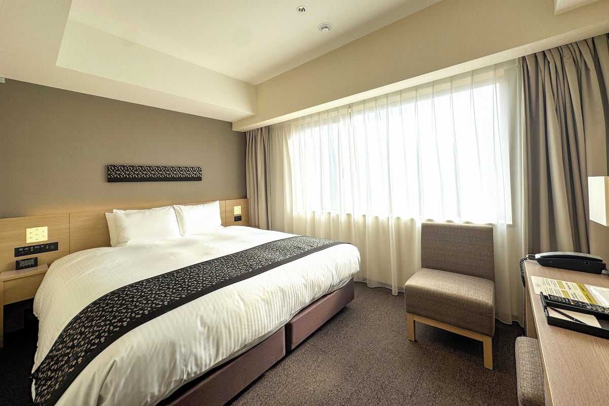 Habitación de hotel con una cama grande, un escritorio con silla, una ventana con cortinas y una obra de arte colgada en la pared.