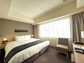 A hotel room with a large bed, a desk with a chair, a window with curtains, and a wall-mounted artwork.