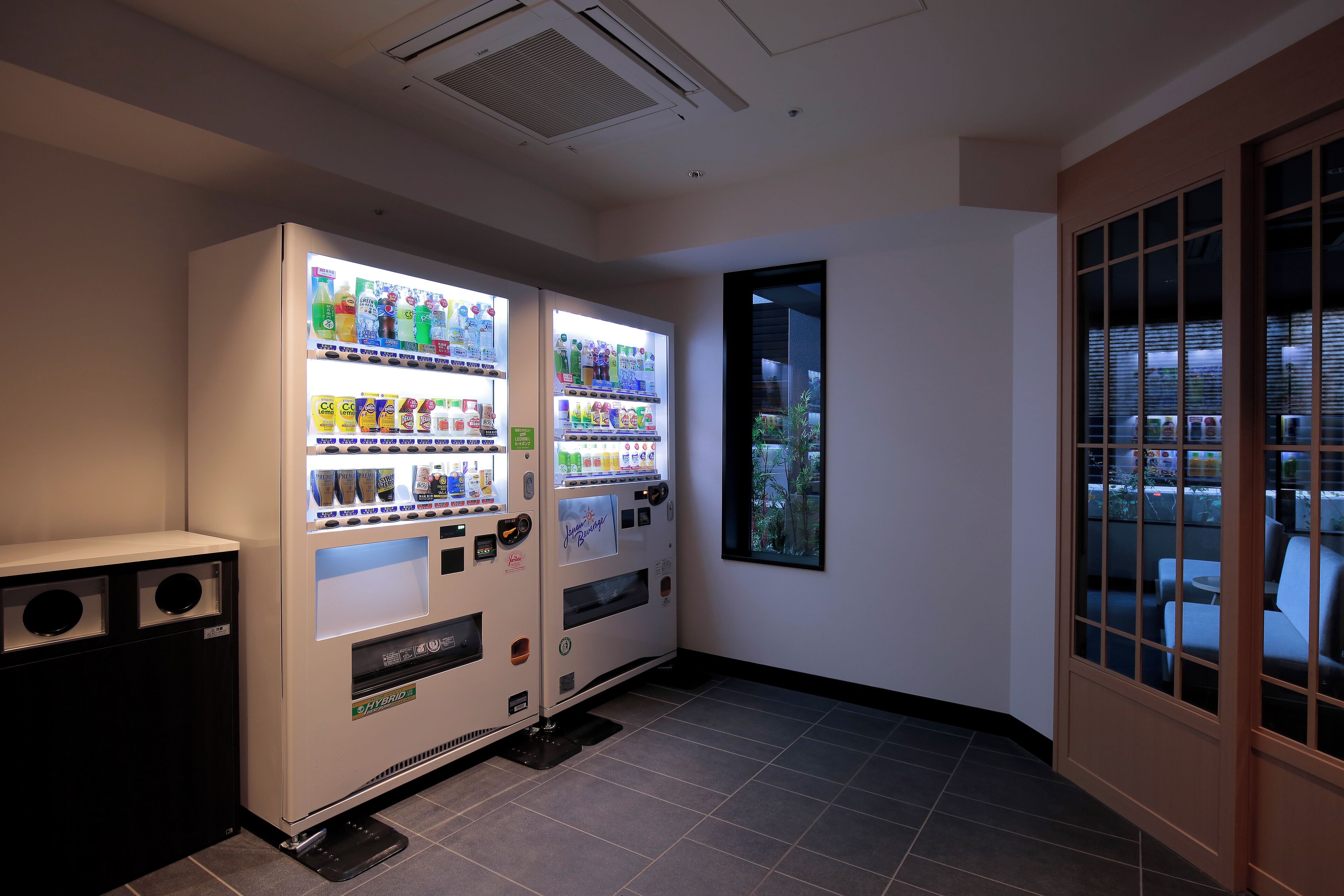 A vending machine area with two machines, a tiled floor, and a view through a window to the outside.