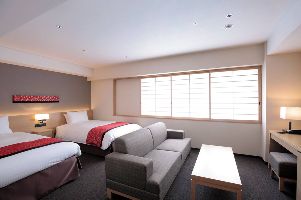 Habitación de hotel con dos camas, un sofá, un escritorio y un ventanal grande.