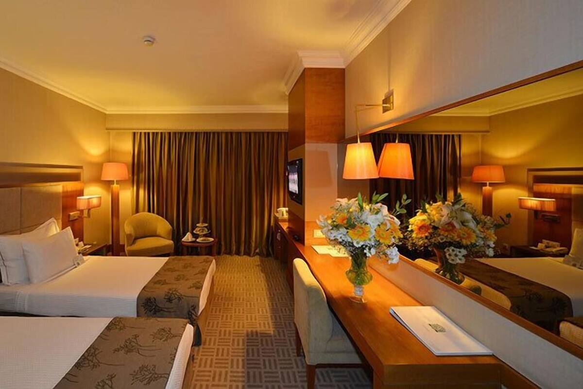 A hotel room with two beds, a desk with a mirror, lamps, a chair, and a vase of flowers.
