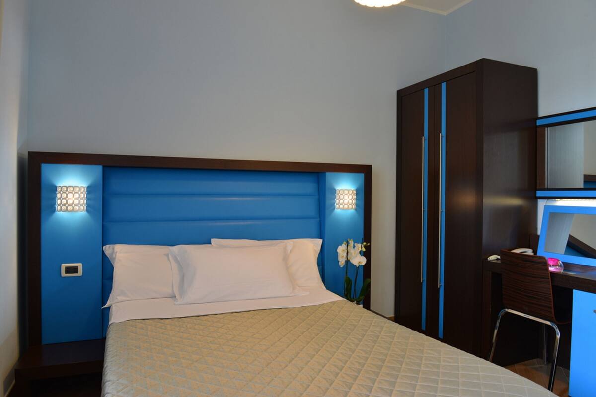 A bedroom with a blue headboard, a bed with white bedding, a desk with a chair, and a mirror.