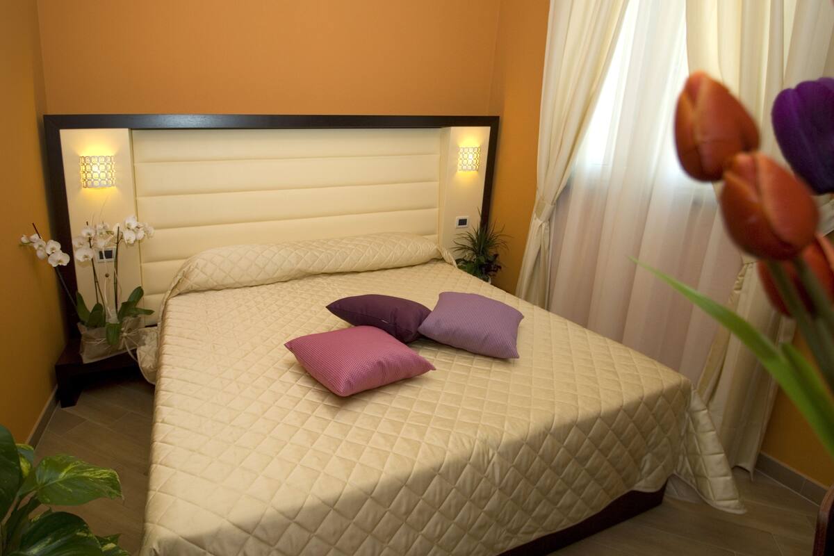 A neatly made bed with a quilted cover and two pillows, a bedside table with a lamp, and a potted plant beside the bed.