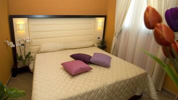 A neatly made bed with a quilted cover and two pillows, a bedside table with a lamp, and a potted plant beside the bed.