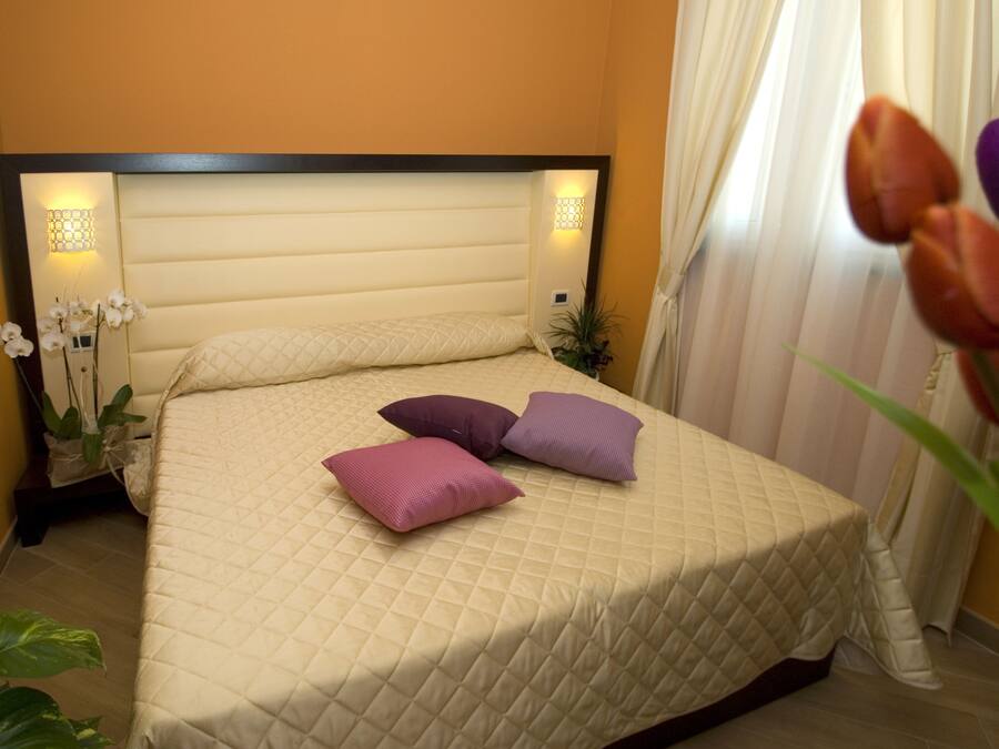 A neatly made bed with a quilted cover and two pillows, a bedside table with a lamp, and a potted plant beside the bed.