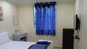A hotel room with a bed, a desk with a chair, a TV mounted on the wall, and a window with blue curtains. - Maha Hotel (Puchong)