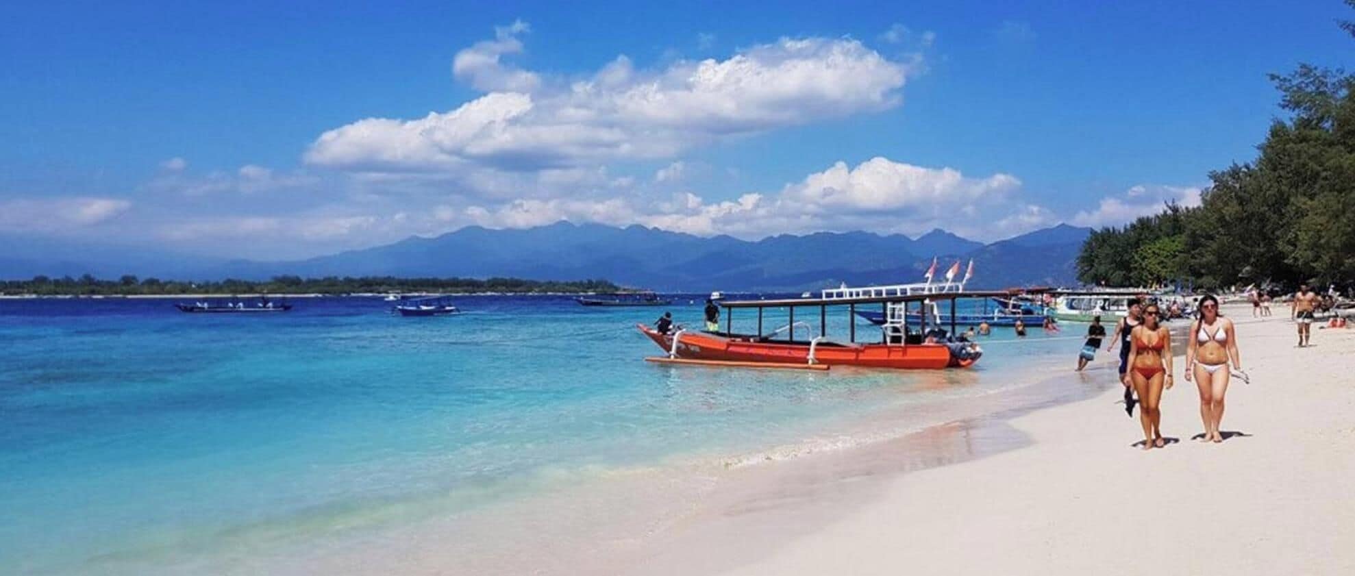 Une plage bordée d’une eau turquoise et limpide, un bateau rouge et, au loin, des montagnes.
