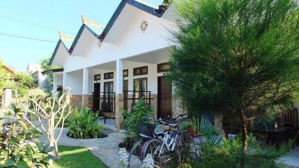 A two-story house with a covered porch, a bicycle parked in front, and a well-maintained garden with various plants and flowers. - Padanta Homestay (Gili Trawangan)