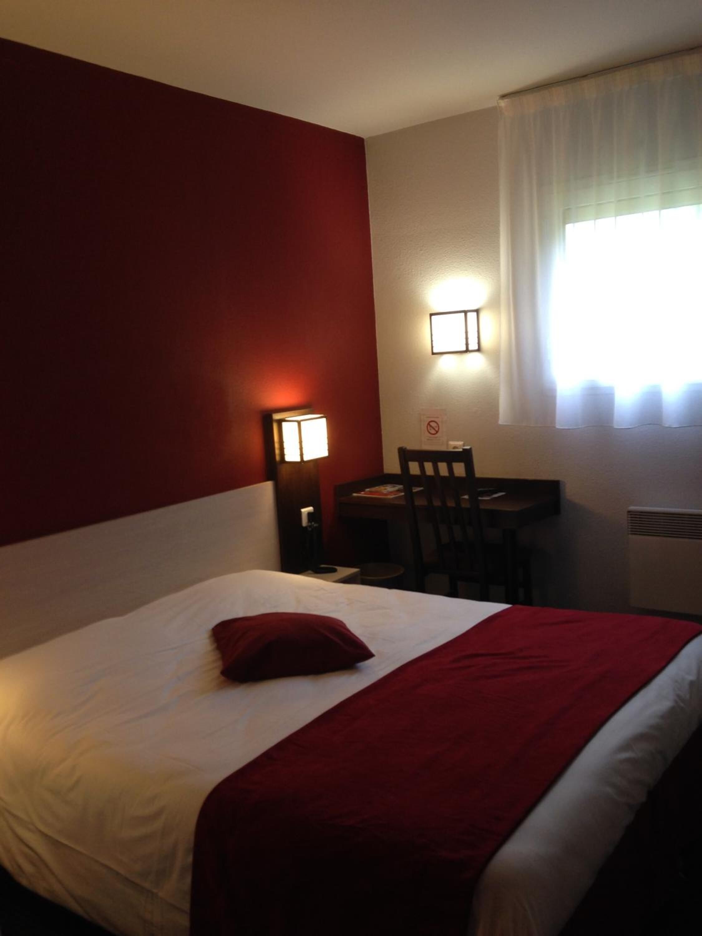 Une chambre d’hôtel avec un lit, un bureau, une chaise, une lampe et une fenêtre avec des rideaux.