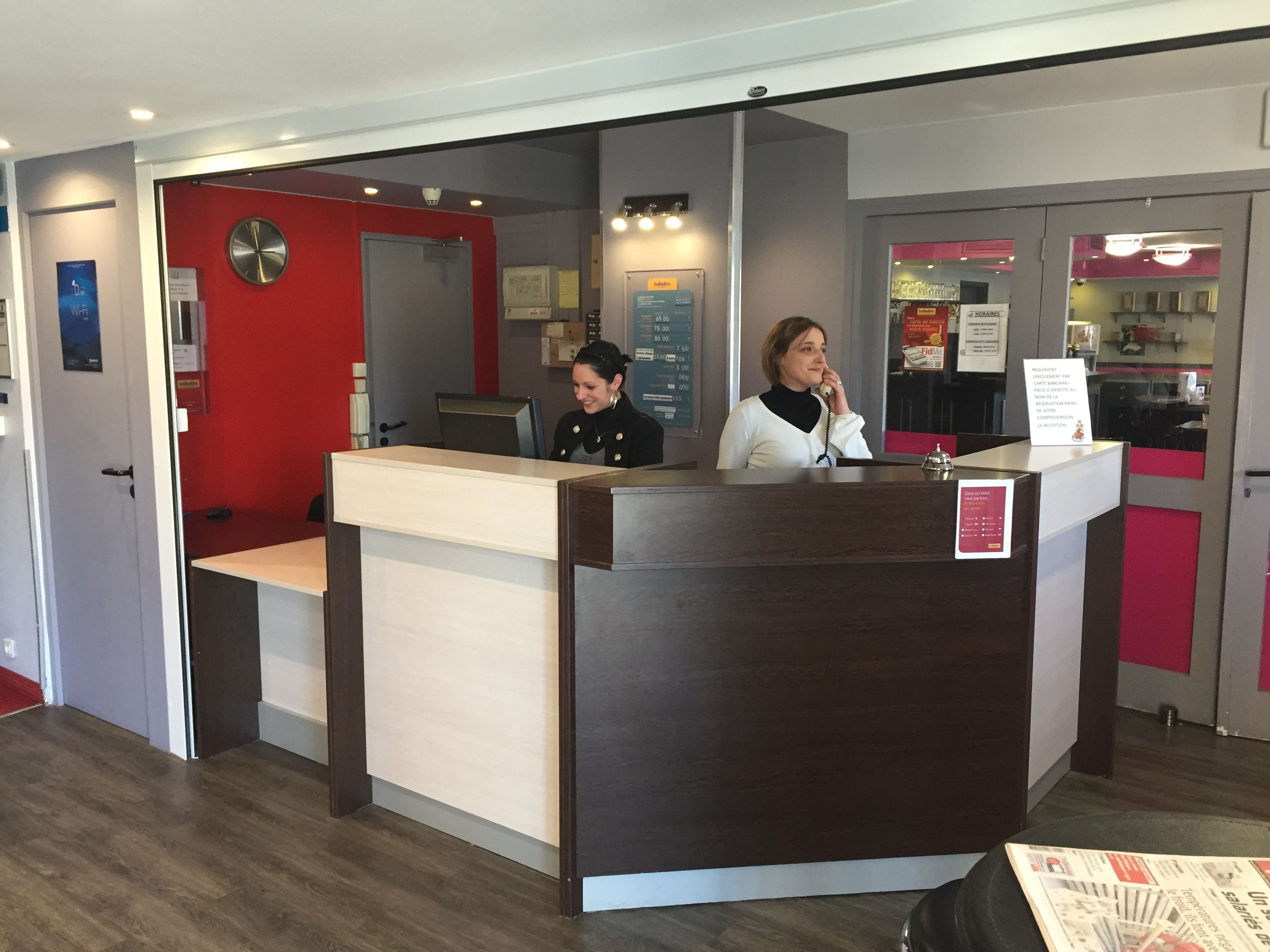 A reception desk with a wooden counter and a red wall behind it. There is a clock on the wall and a menu board to the right.