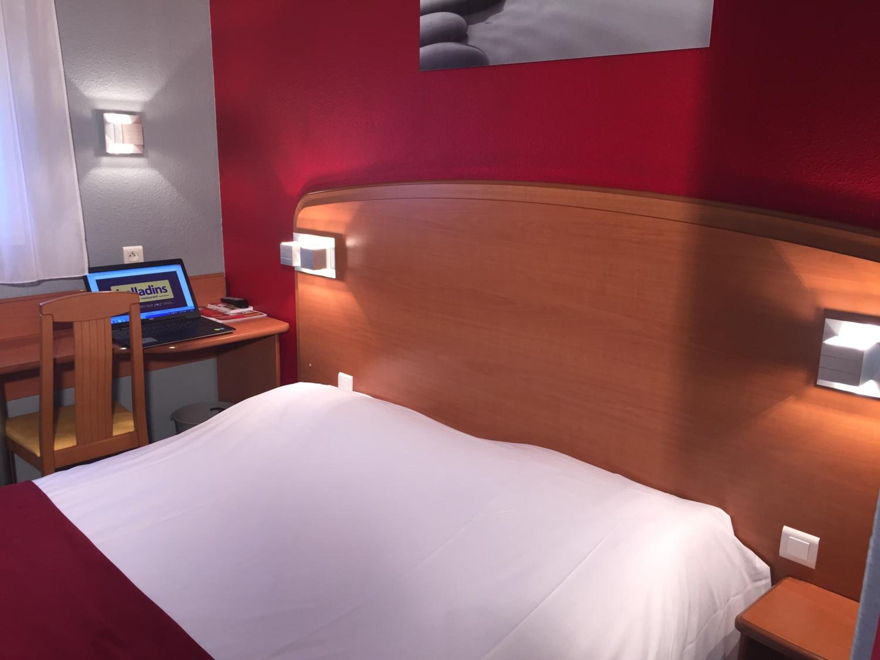 A hotel room with a wooden headboard, a bed with white bedding, a desk with a laptop, and a chair.