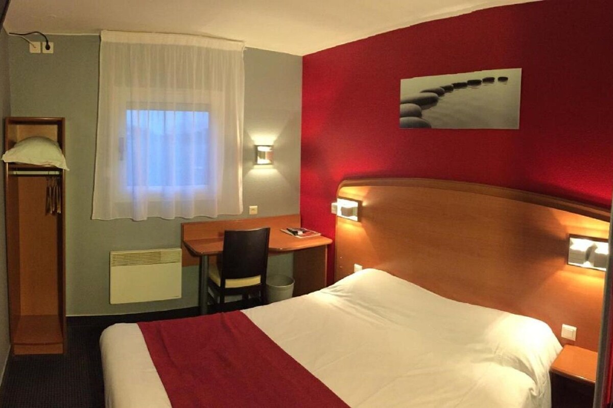 Habitación de hotel con cama, escritorio, silla, ventana con cortinas, lámpara de pared y un cuadro.
