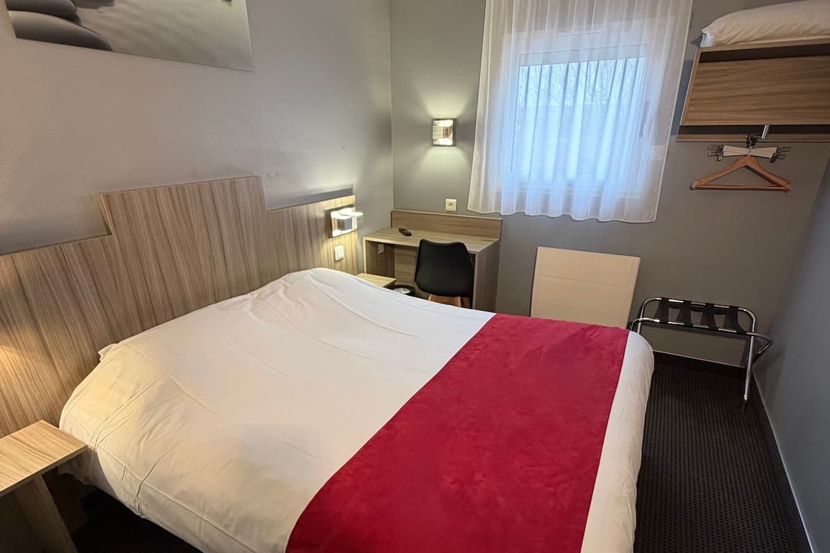 Habitación de hotel con cama, escritorio, silla, perchero y ventana con cortinas.