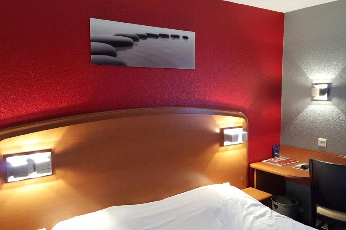 Habitación de hotel con cabecera de madera, dos lámparas de noche, un escritorio pequeño con una silla y una obra de arte colgada en la pared.