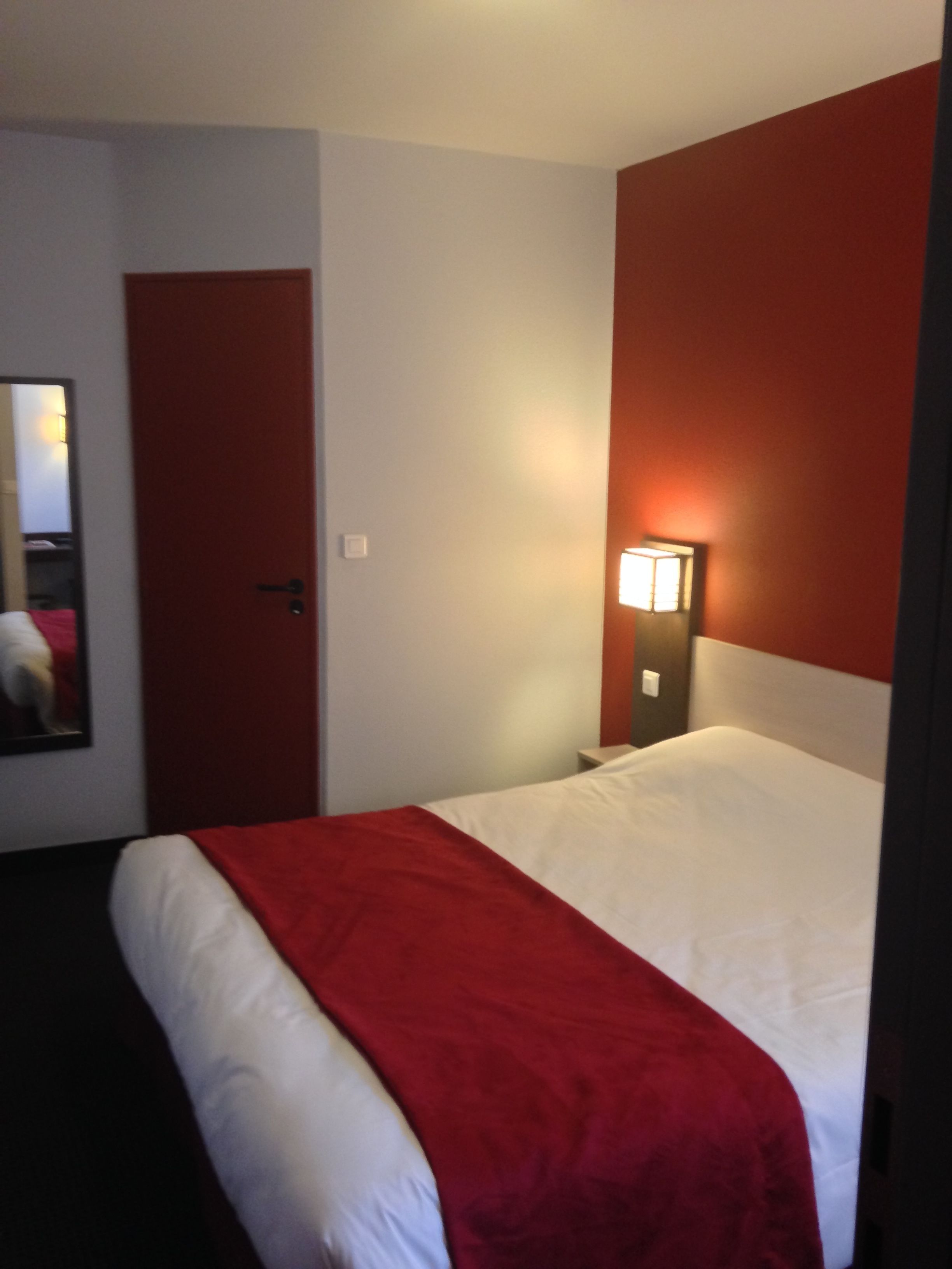 Habitación de hotel con una cama, pared roja, pared blanca, puerta, mesita de noche y lámpara fijada a la pared.