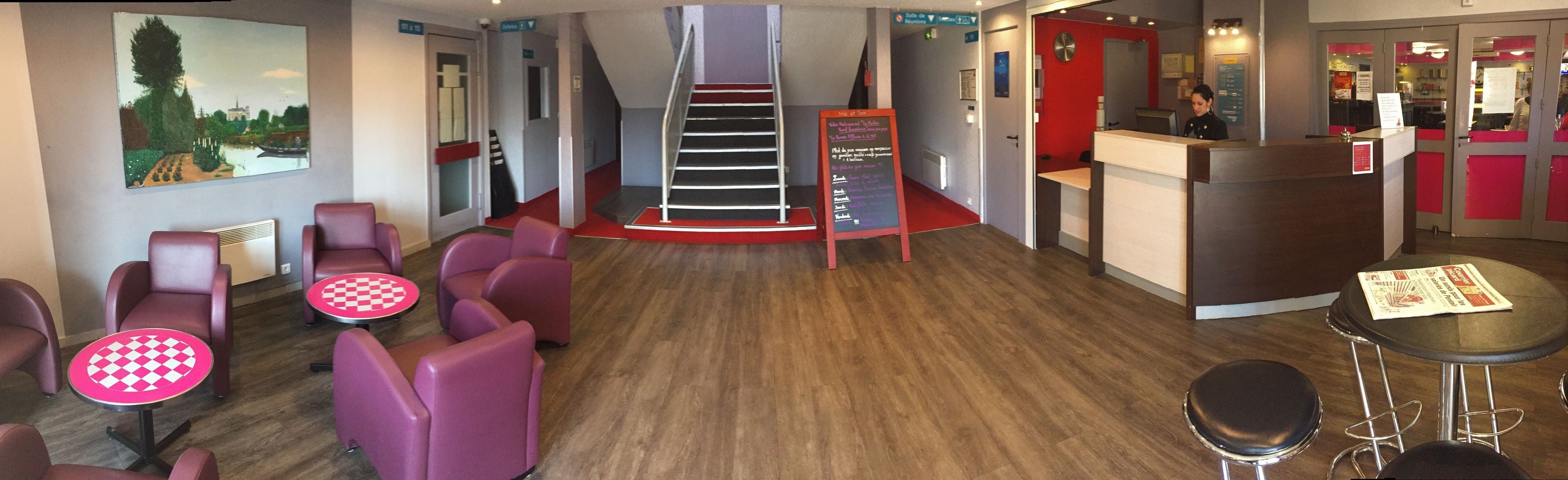 Eine Hotelhalle mit einer Rezeption, einer Sitzecke mit Stühlen und Tischen, einer Treppe und einem Wandbild.
