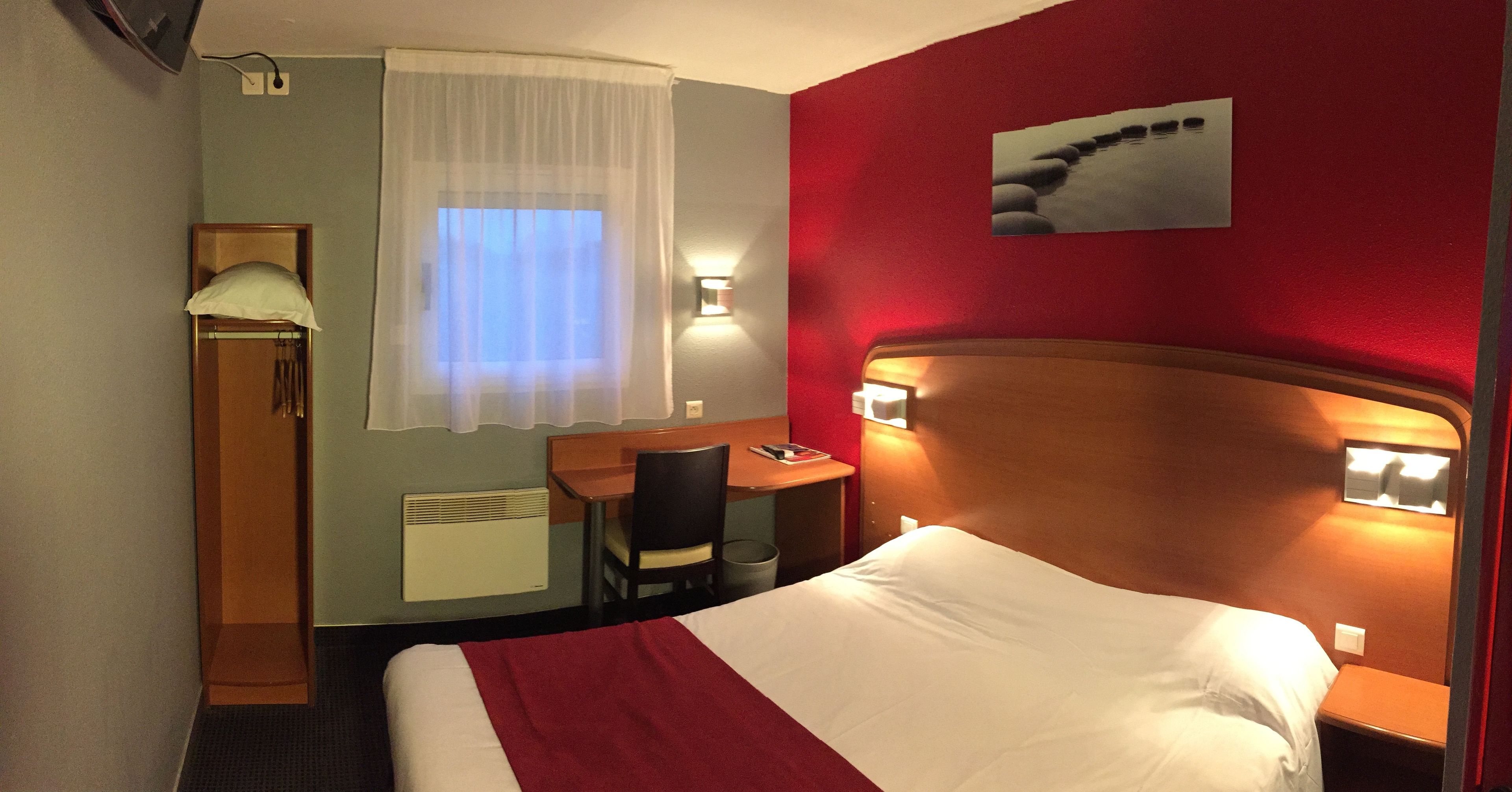 A hotel room with a bed, a desk, a chair, a window with curtains, a wall-mounted picture, and a wall-mounted lamp.