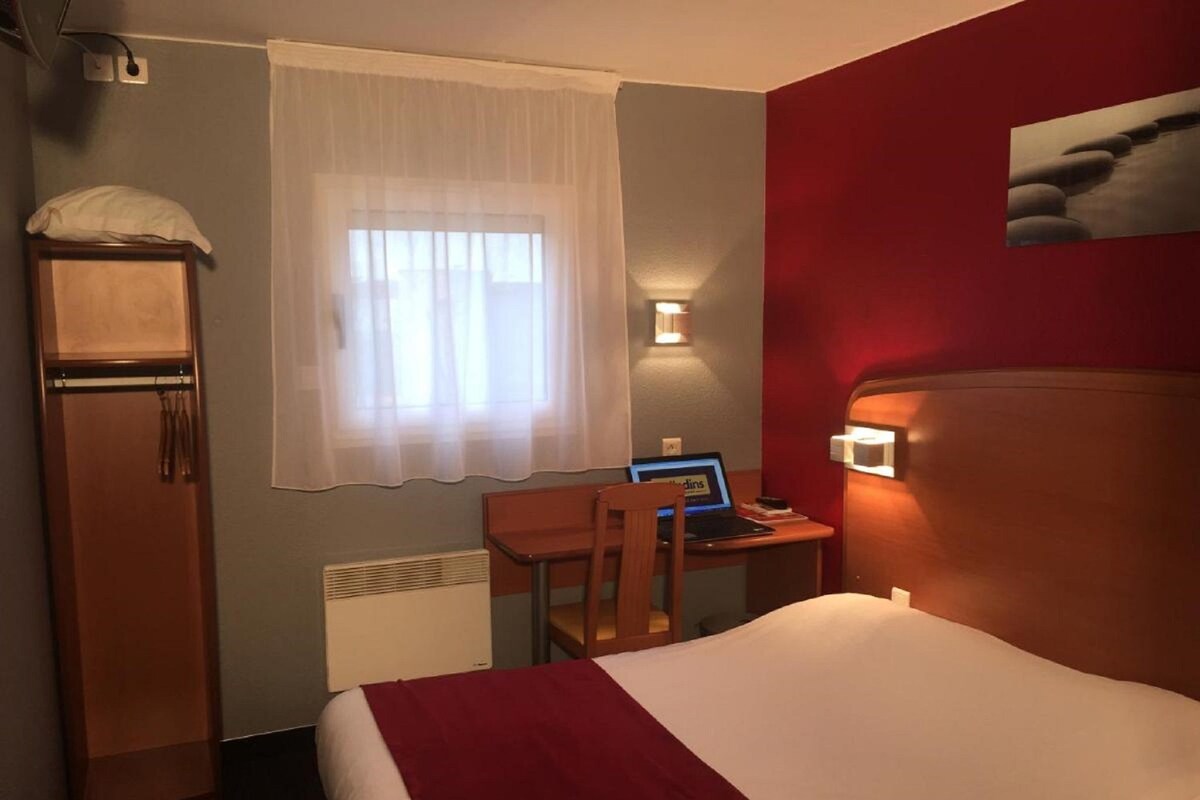 Een hotelkamer met een bed, een bureau met een laptop, een stoel, een kledingkast, een radiator en een raam met gordijnen.
