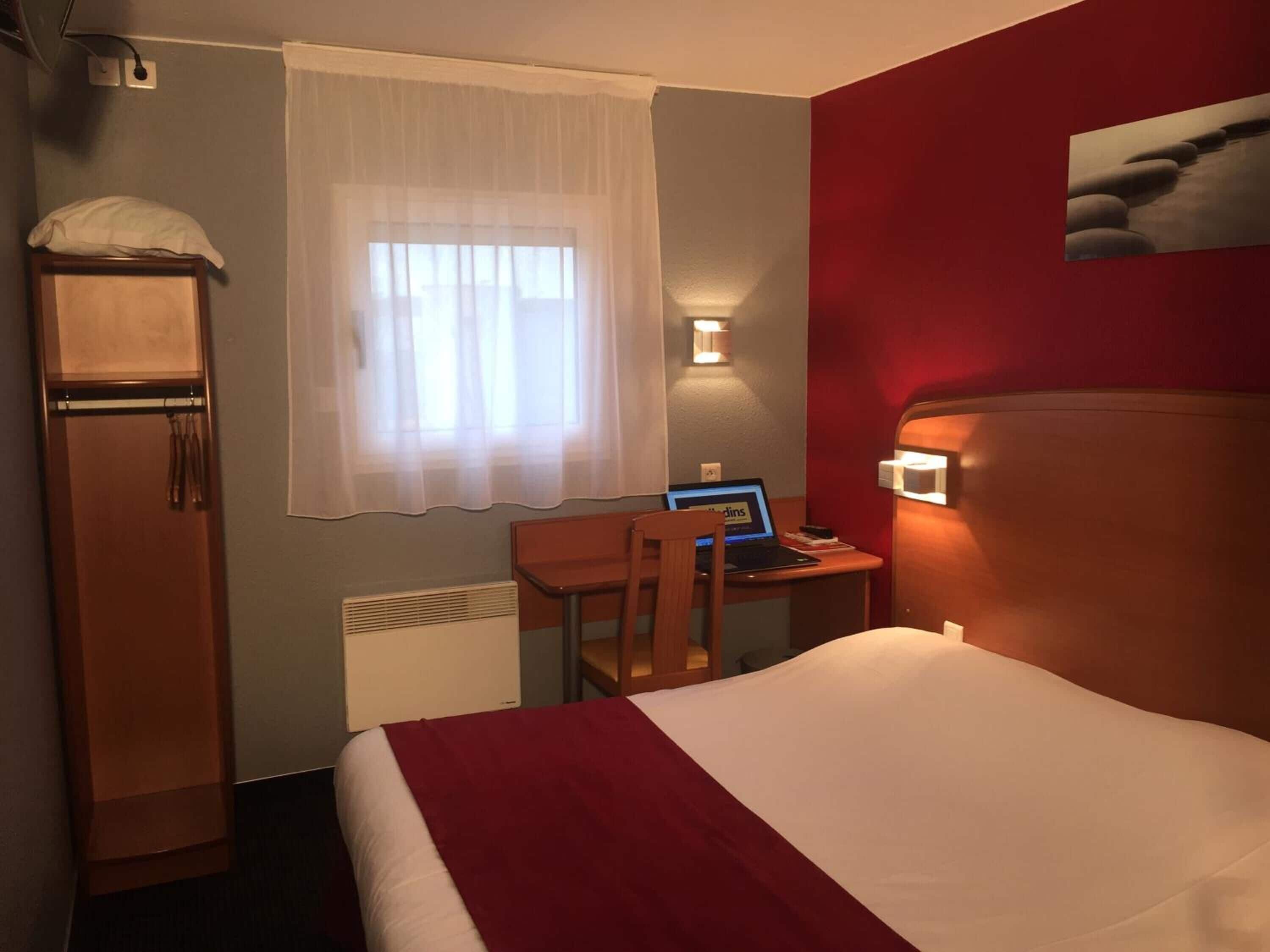 A hotel room with a bed, a desk with a laptop, a chair, a wardrobe, a window with curtains, and a wall-mounted light.