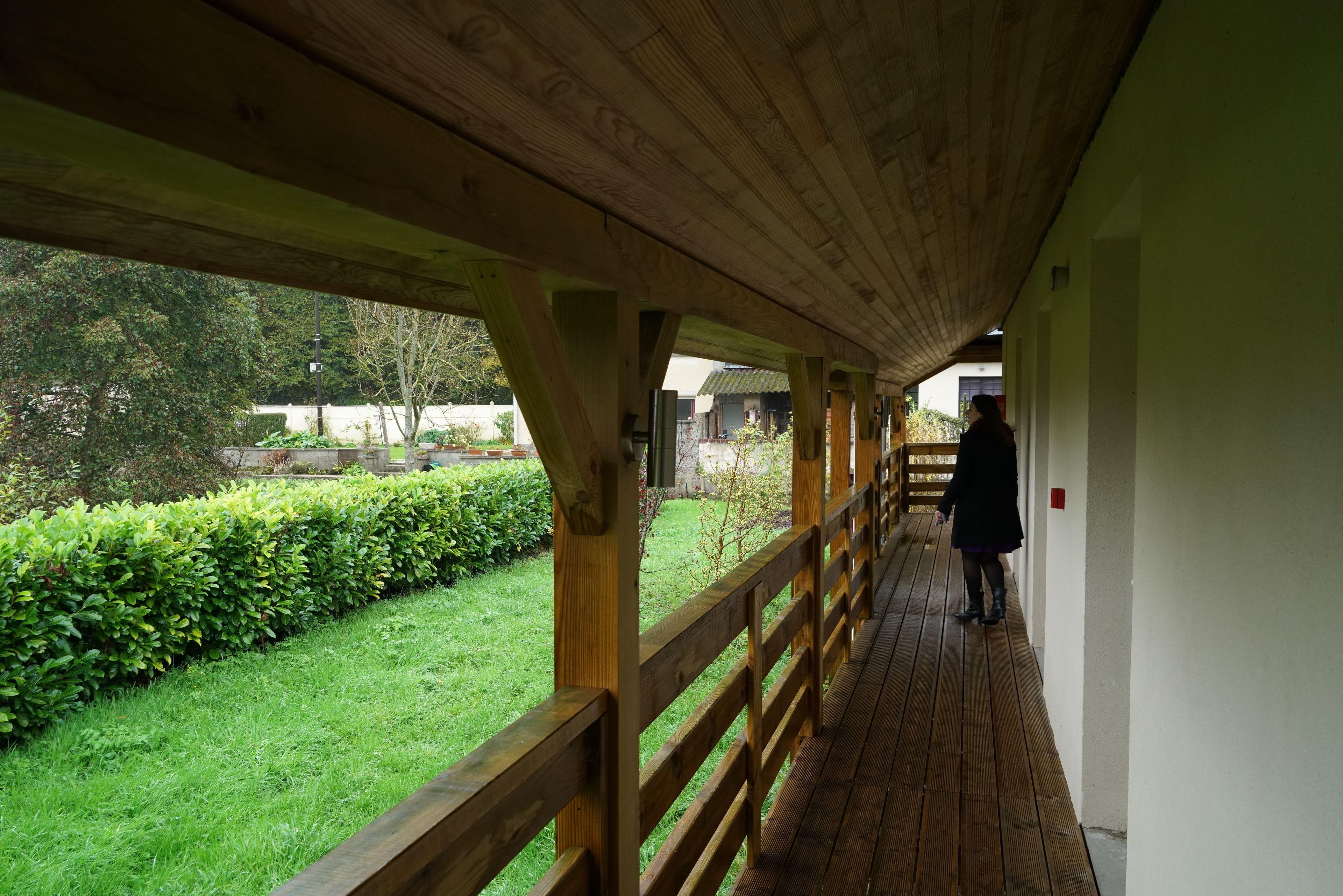 A covered wooden walkway with a person walking away, bordered by a hedge and a grassy area.