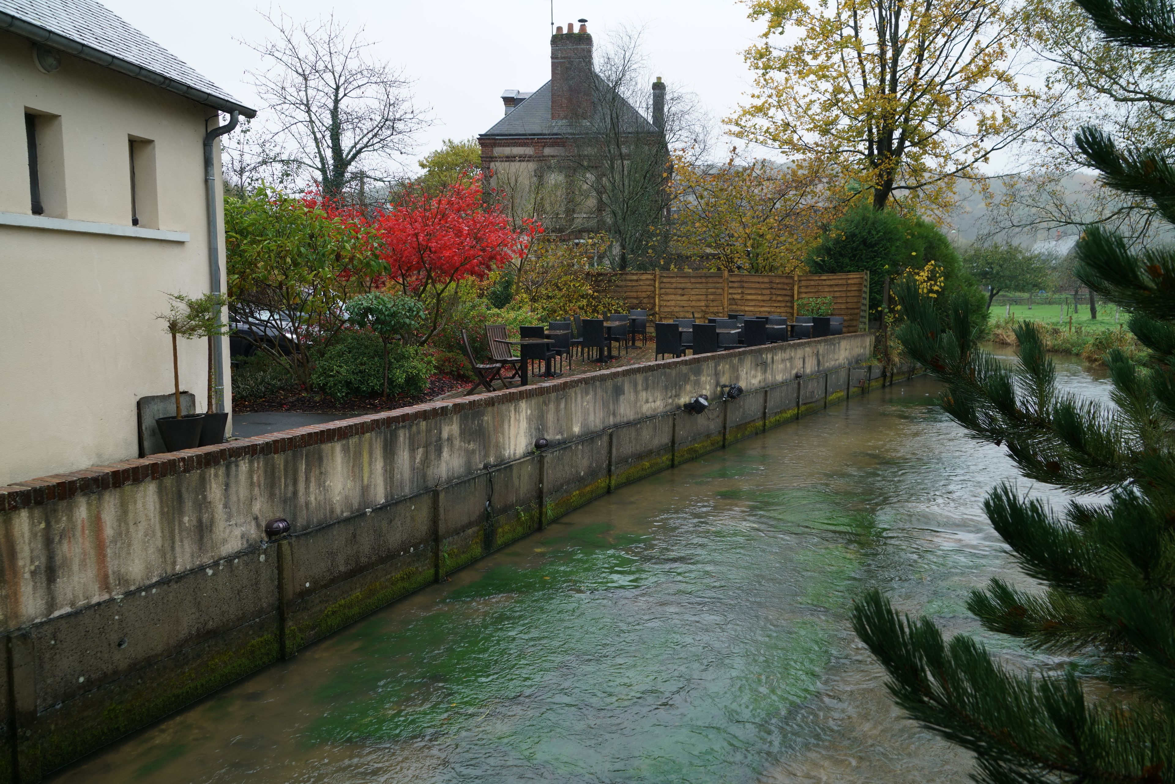 A riverside walkway with outdoor seating, a stone wall, and a building in the background.