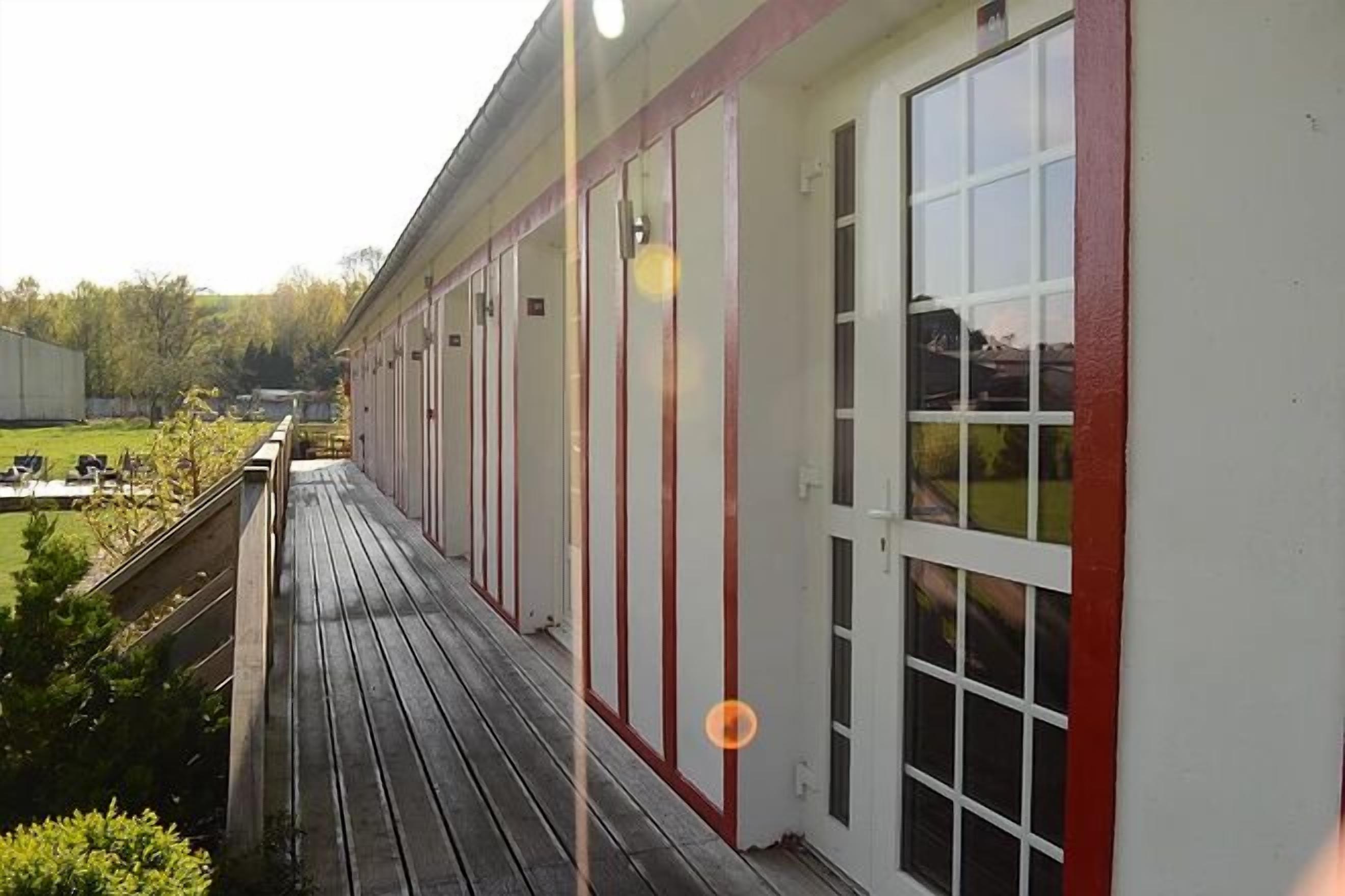 A building with a long, narrow corridor with large windows and doors, a wooden deck in front, and a grassy area in the background.