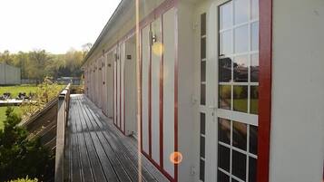 A building with a long, narrow corridor with large windows and doors, a wooden deck in front, and a grassy area in the background.
