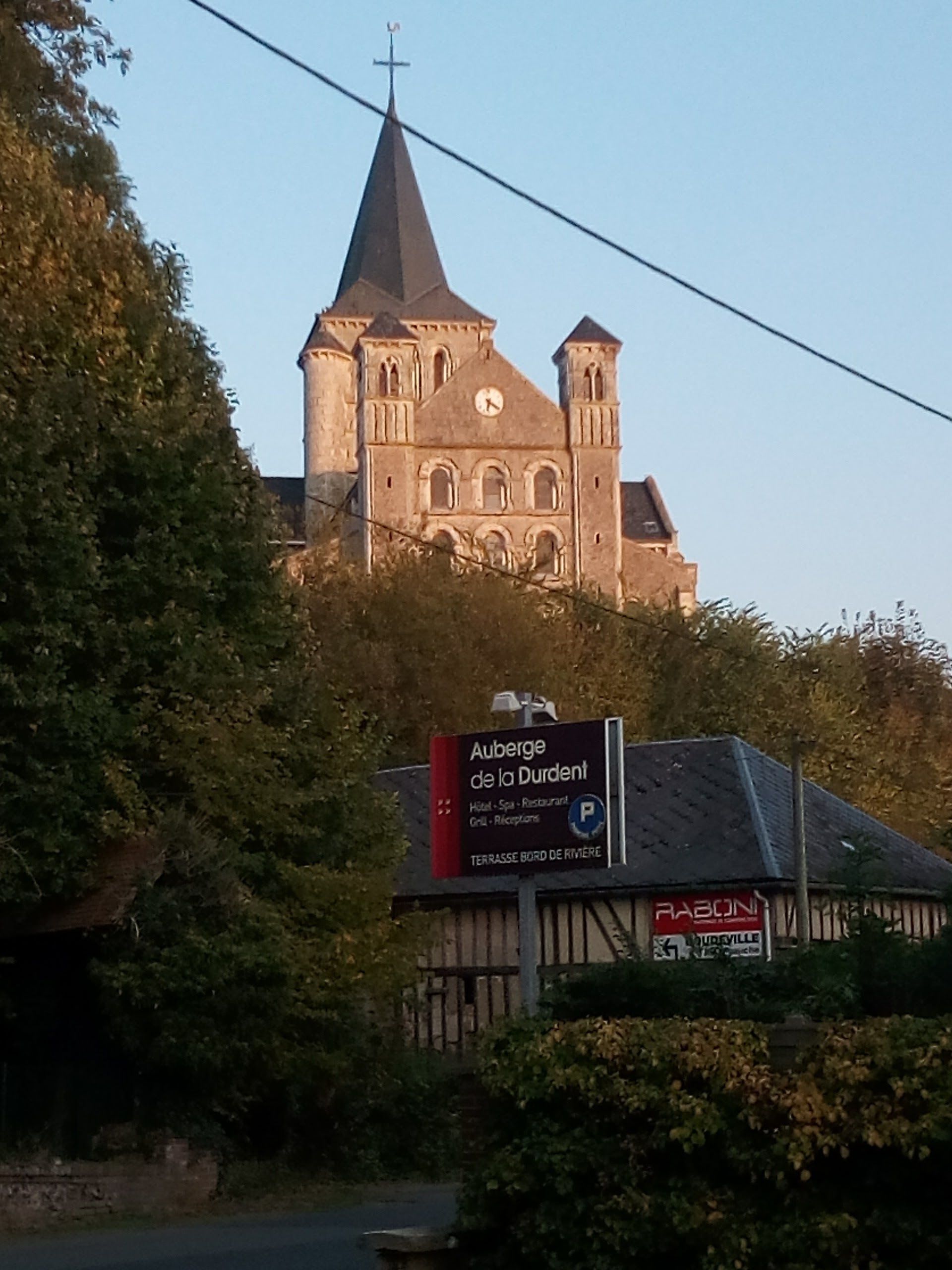 Una iglesia con torre del reloj, rodeada de árboles y un cartel de Auberge de la Durent.