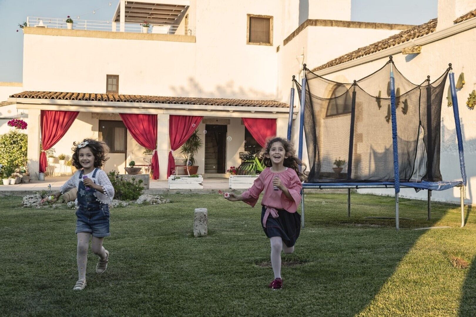Zwei Kinder spielen in der Nähe eines Trampolins auf einer grünen Wiese, im Hintergrund ist ein Haus zu sehen.