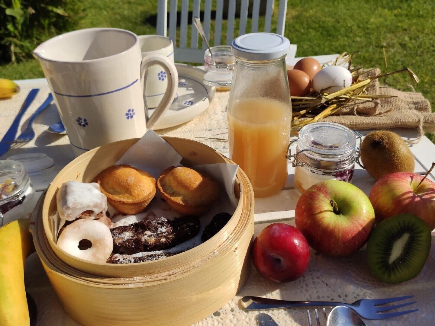 A picnic setup with food items, including a basket of pastries, fruits, and a glass of juice on a table outdoors.