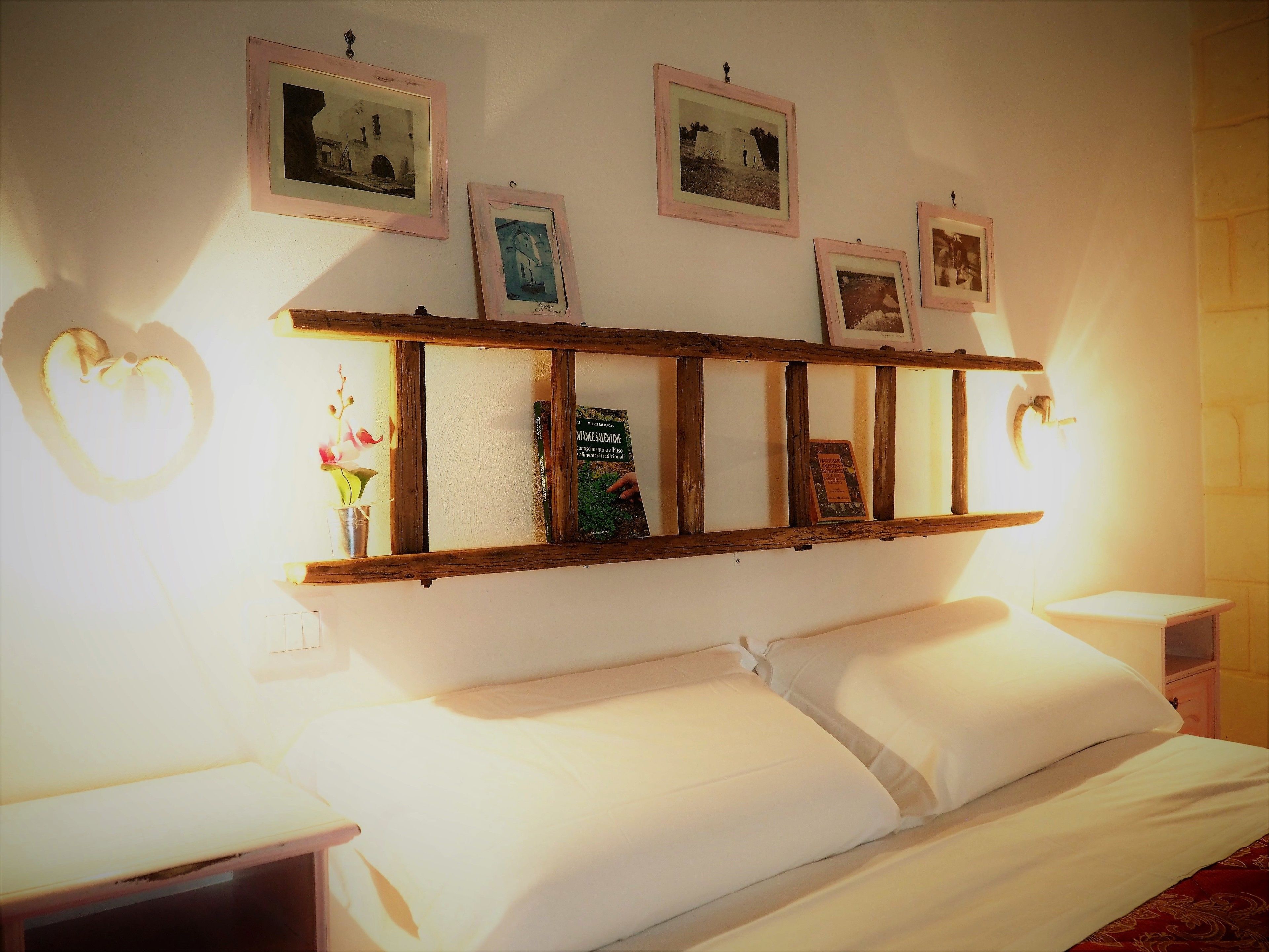 A neatly made bed with white linens, a wooden shelf above holding framed pictures and a book, and a heart-shaped wall decoration.