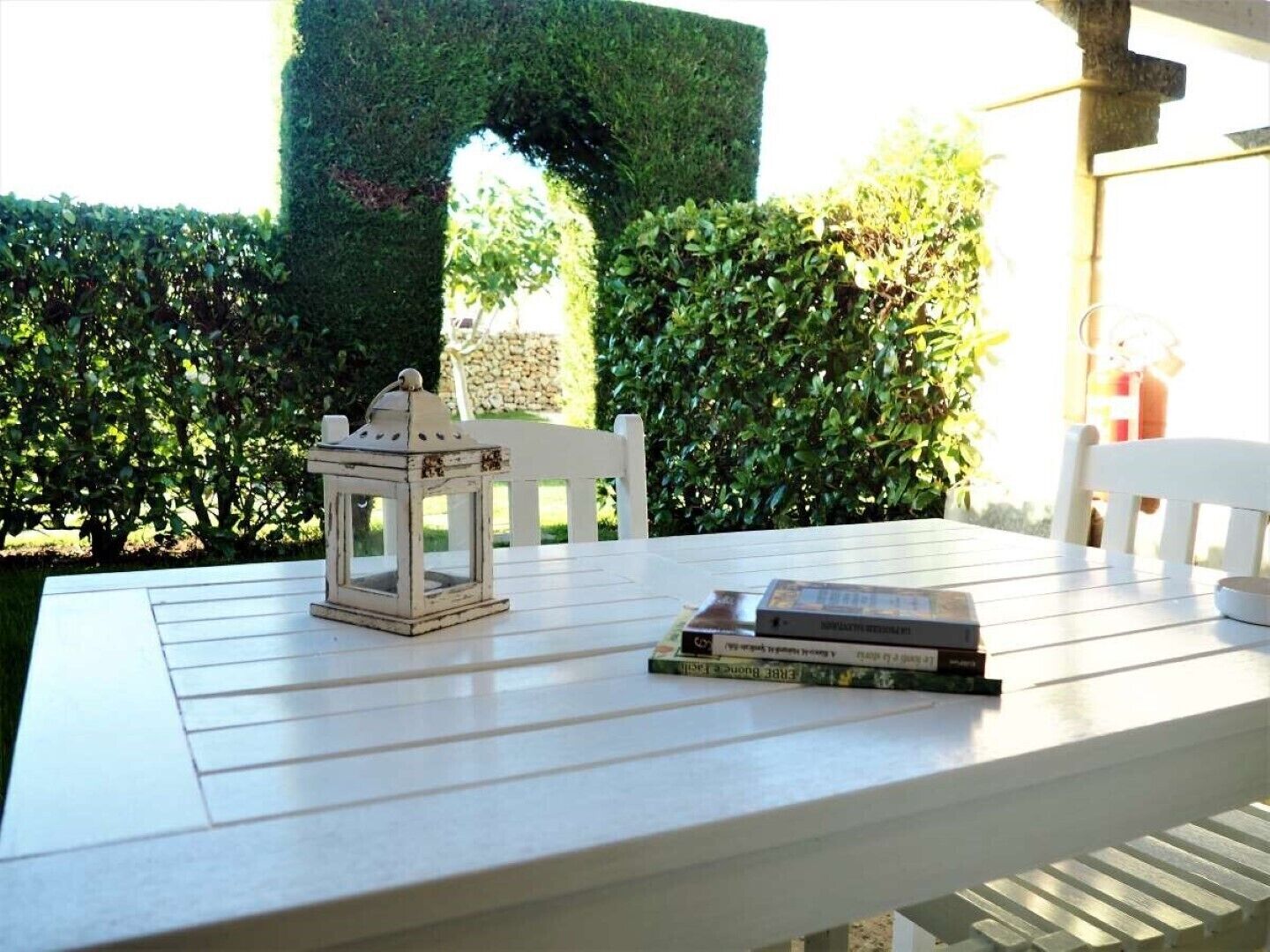 Una mesa blanca con un farol y libros, frente a un fondo de setos ordenados y una estructura de piedra.