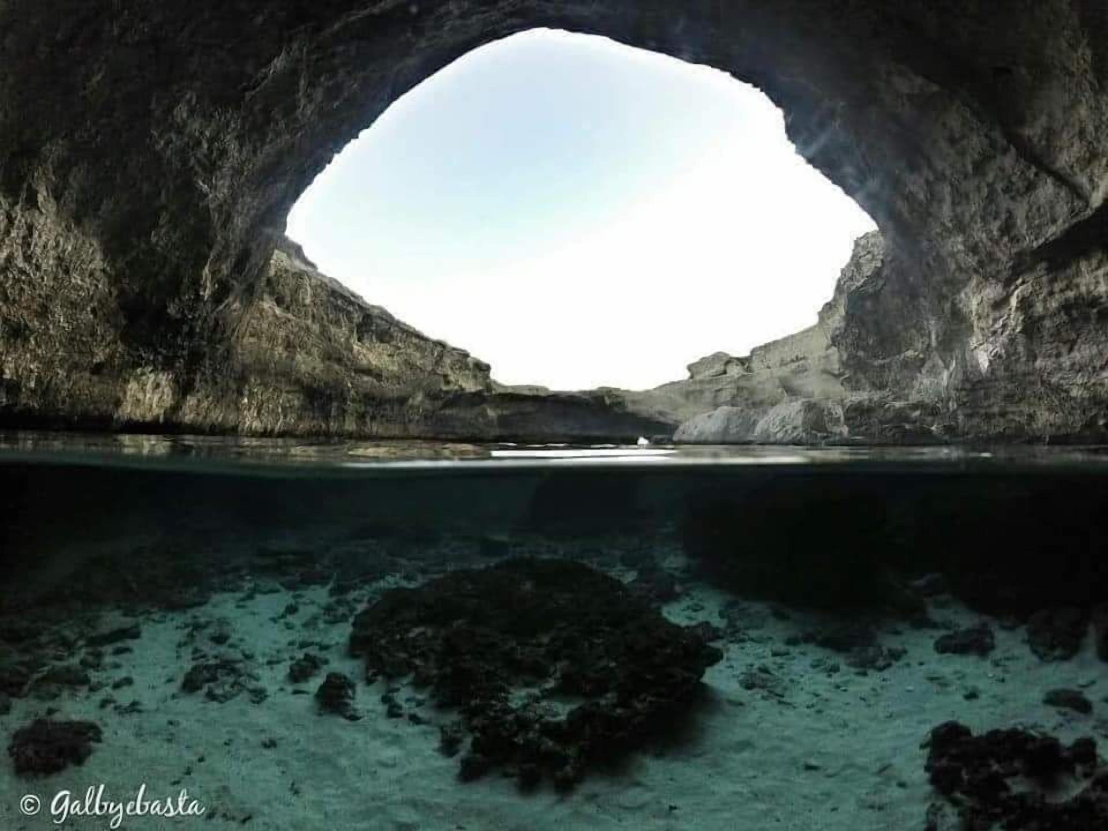 Boca natural de cueva con vista a una masa de agua tranquila y formaciones rocosas.