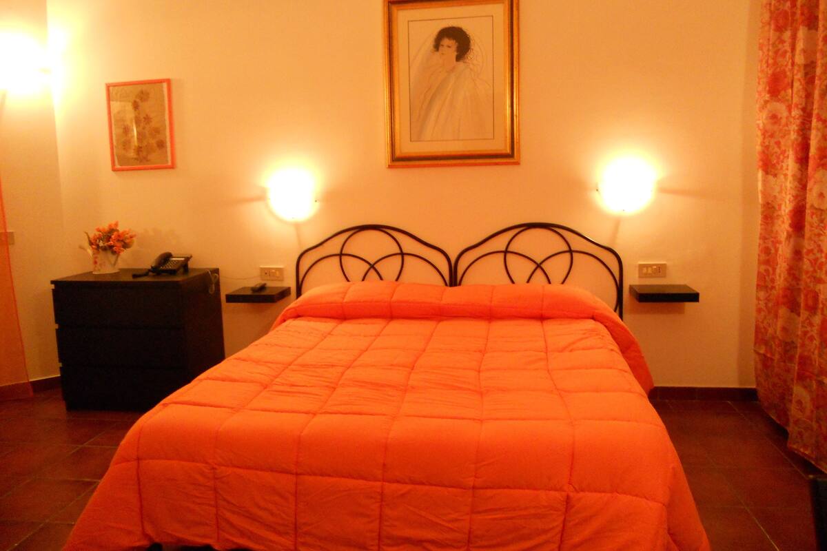 Ein Schlafzimmer mit einem Bett, zwei Nachttischlampen, einem Nachttisch mit Telefon, einem gerahmten Bild an der Wand und einem blumigen Vorhang.