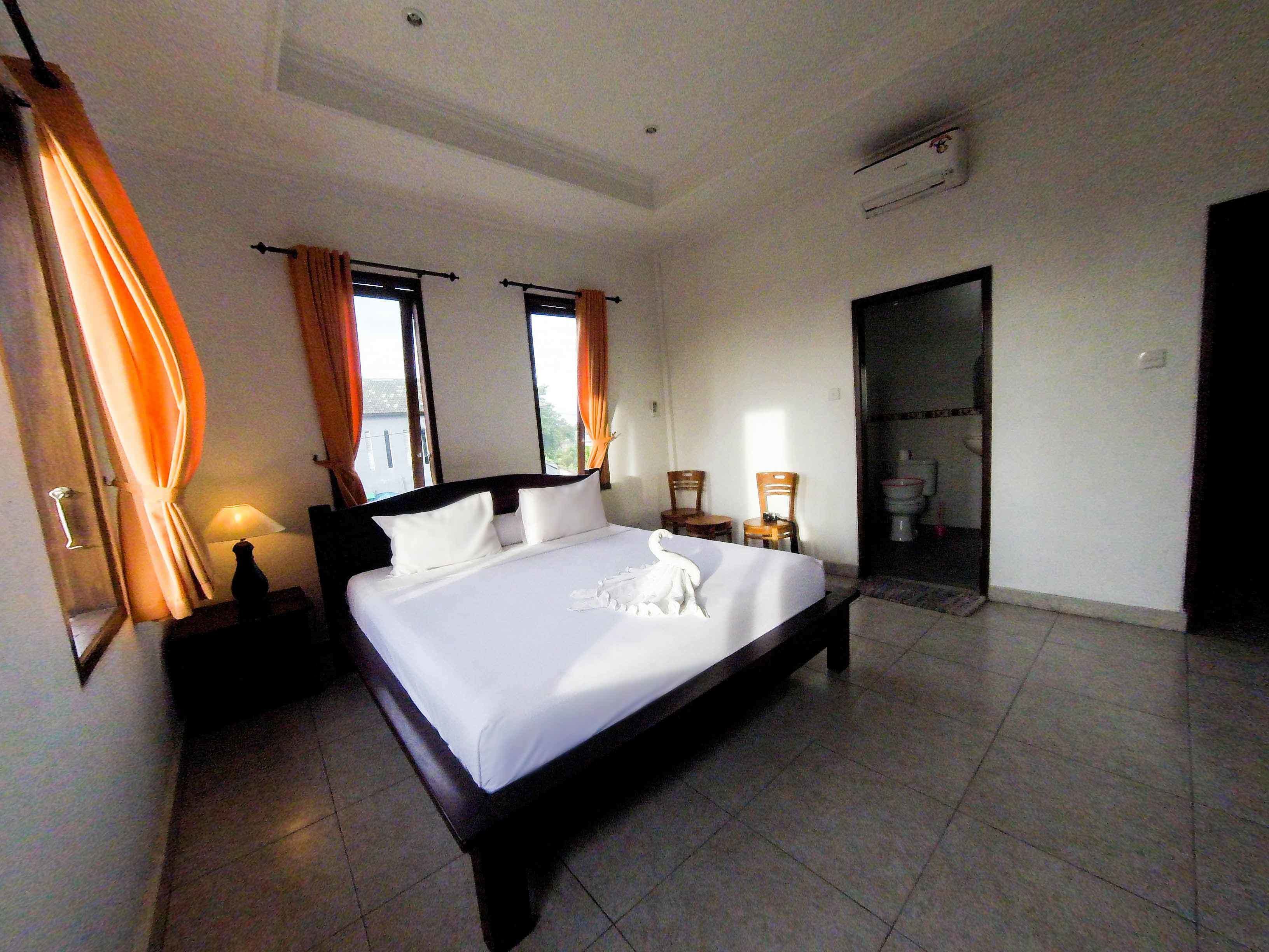 Una cama bien hecha en una habitación de hotel, con un ventanal grande, una mesita de noche con lámpara, y una puerta que da a un baño.