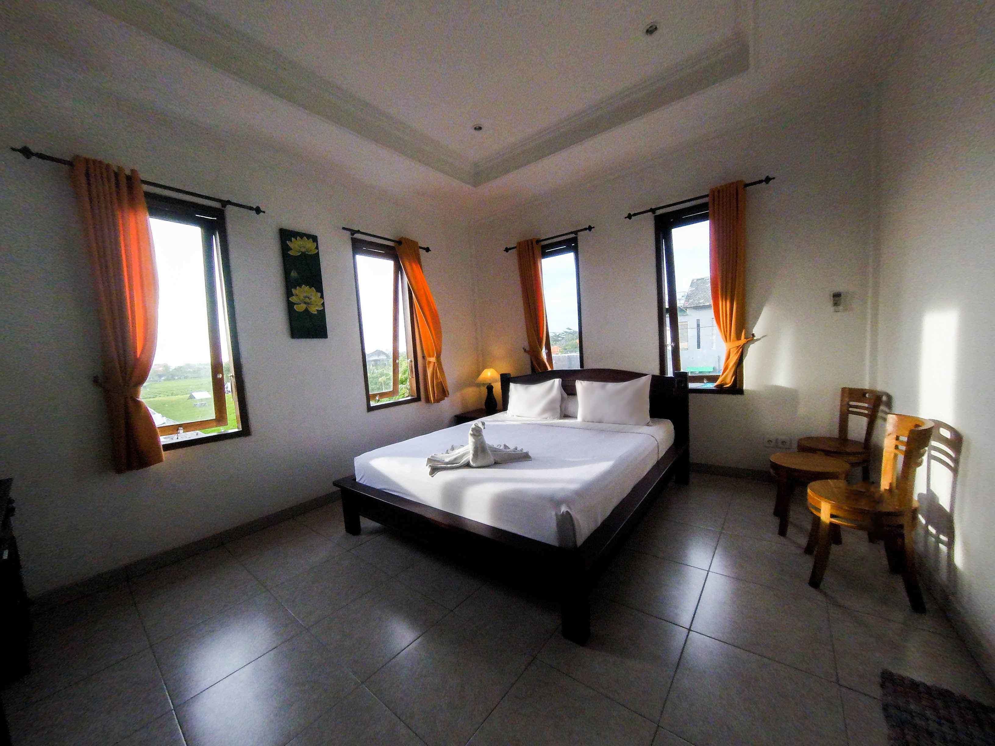 A bedroom with a large bed, two windows with curtains, a small table with chairs, and a lamp.