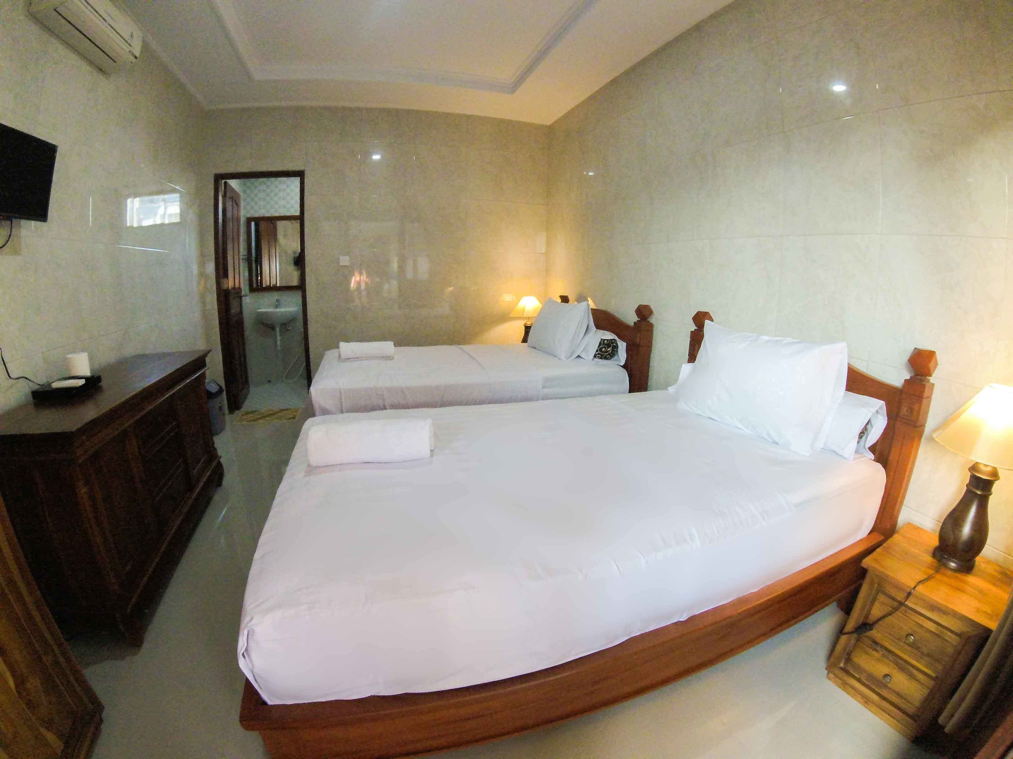 A hotel room with two single beds, a wooden nightstand with a lamp, a wooden dresser, a TV mounted on the wall, and a bathroom with a sink and mirror.