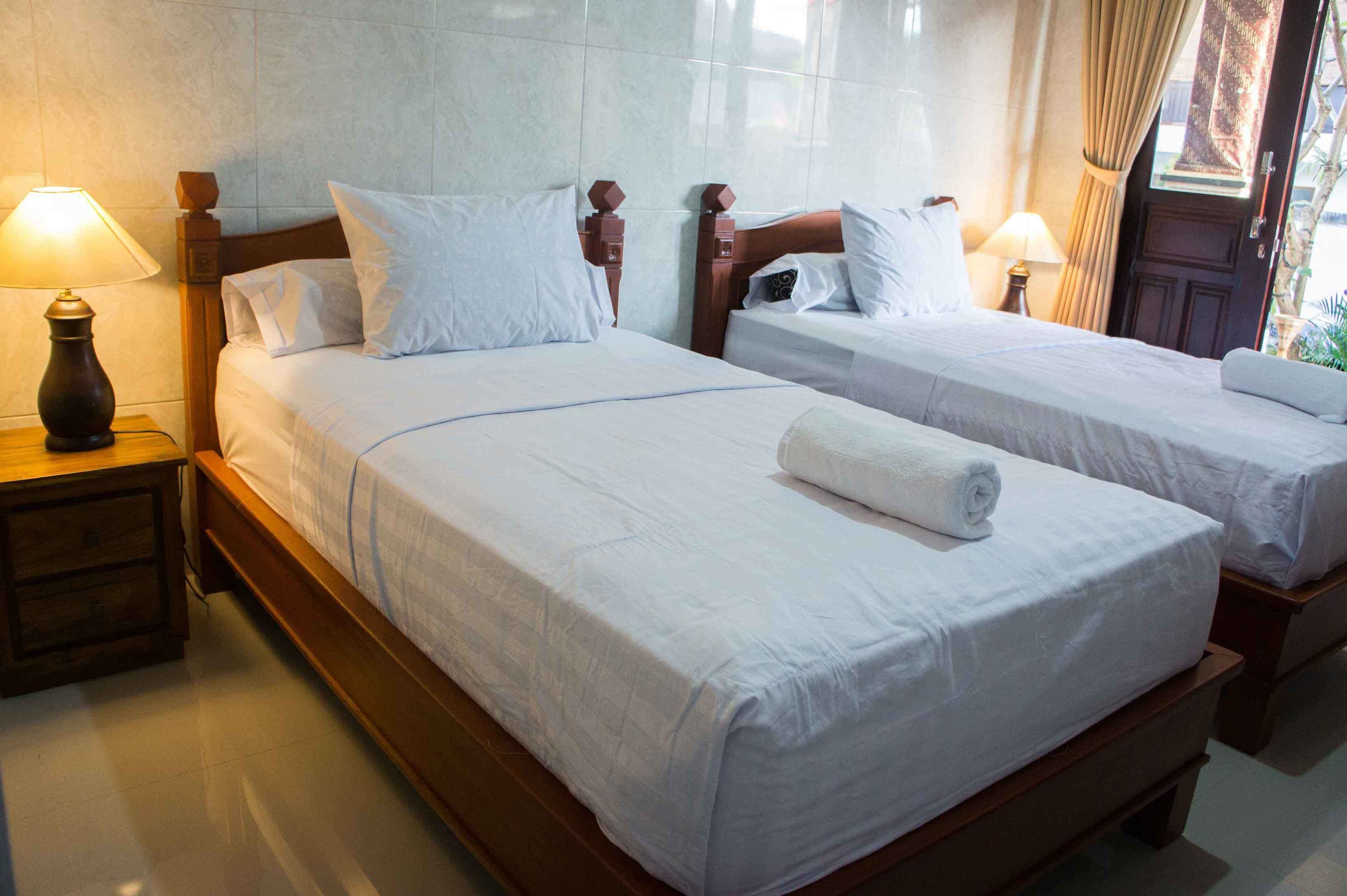 A hotel room with two single beds, wooden headboards, white linens, and bedside lamps.
