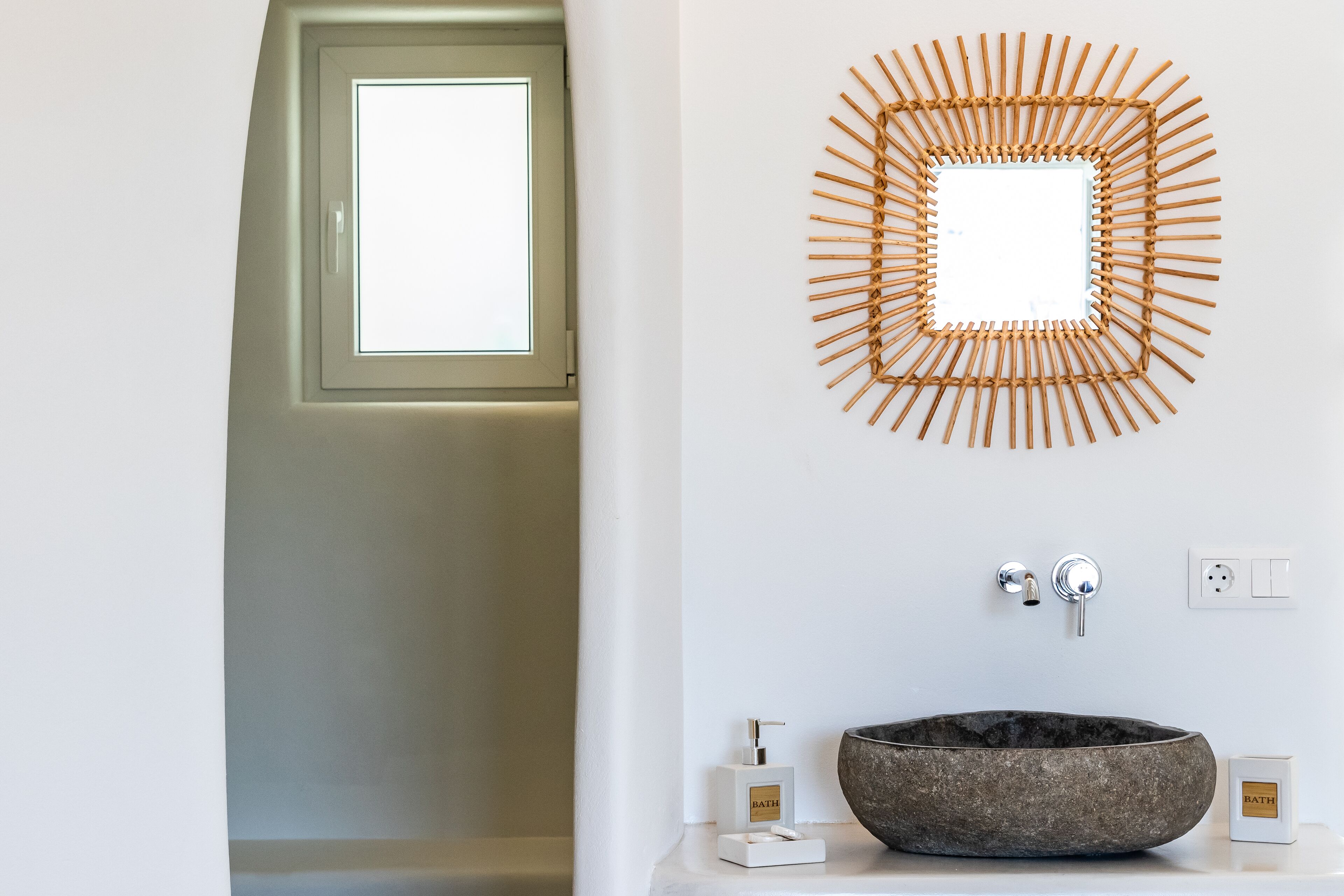 Une salle de bain avec un grand miroir rond en osier, un lavabo en pierre et une fenêtre.
